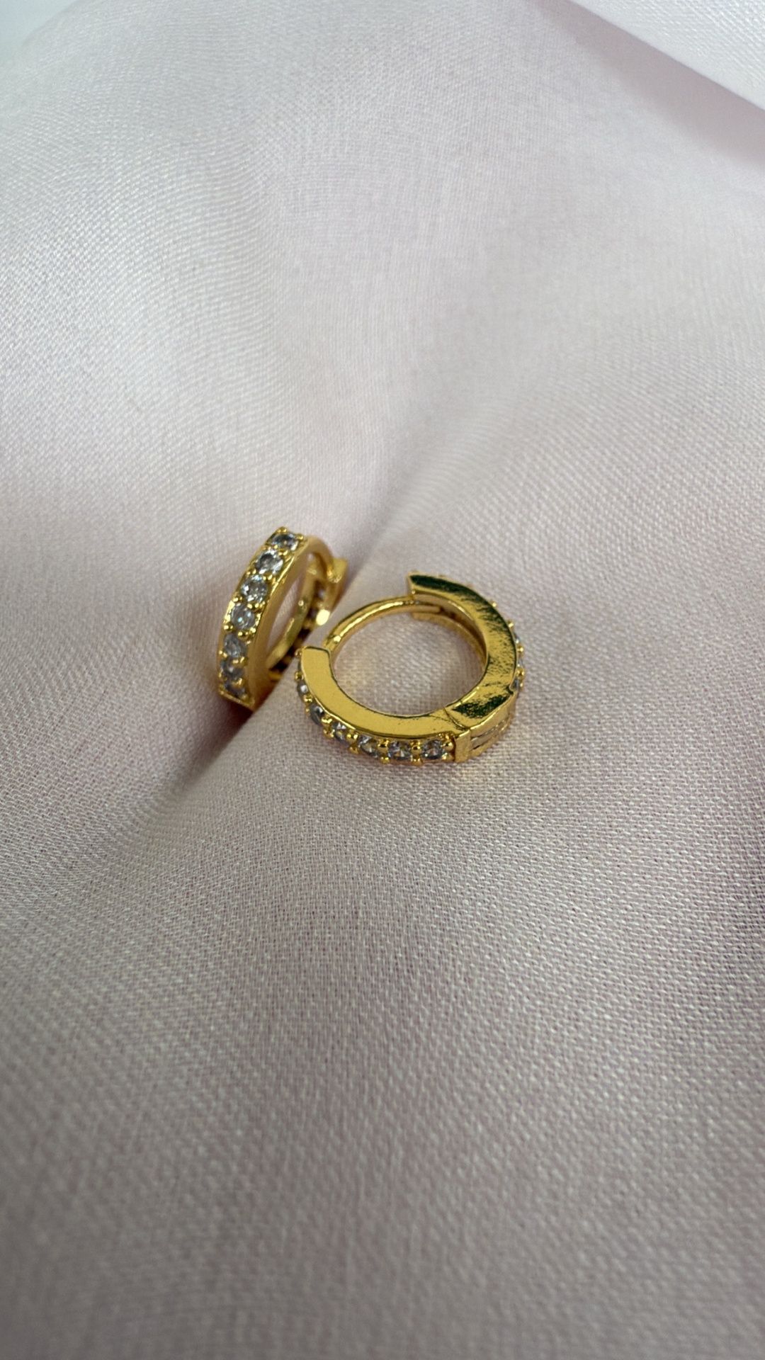 Brincos de argola dourados com pedras brilhantes sobre tecido creme