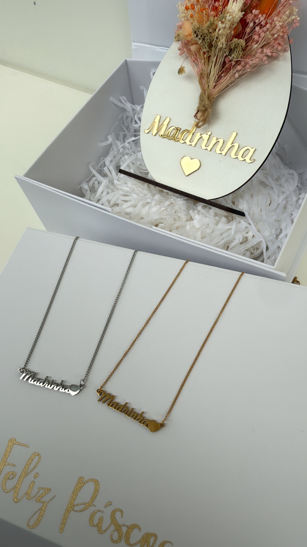 Colares prateados e dourados com pendente Madrinha junto a flores secas e caixa Feliz Páscoa
