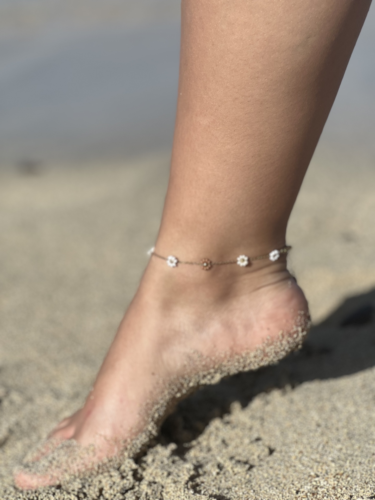 Tornozeleira com flores pequenas num pé na praia, parcialmente coberto de areia