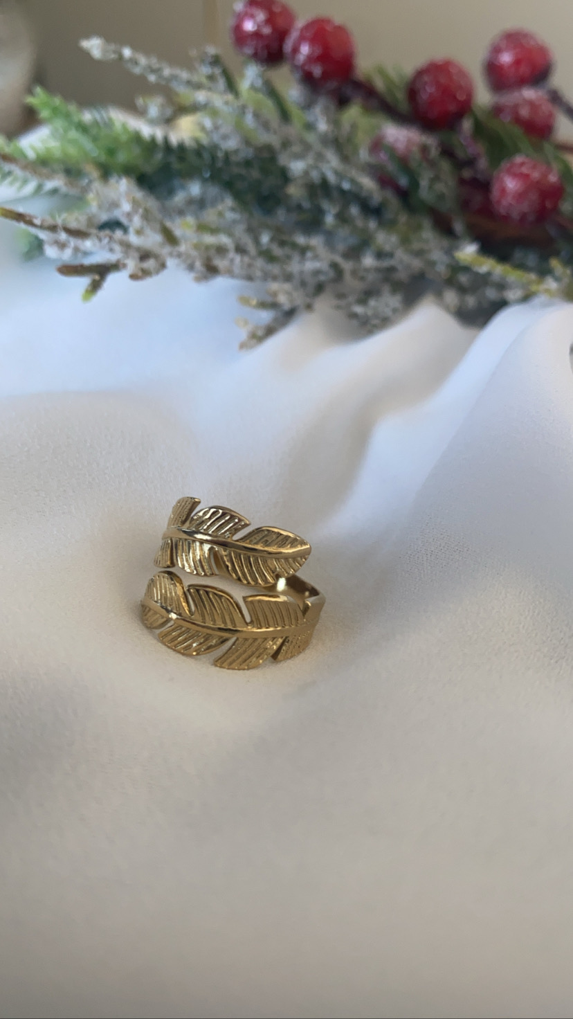 Anel dourado com design de folhas sobre tecido branco e ramos decorativos ao fundo