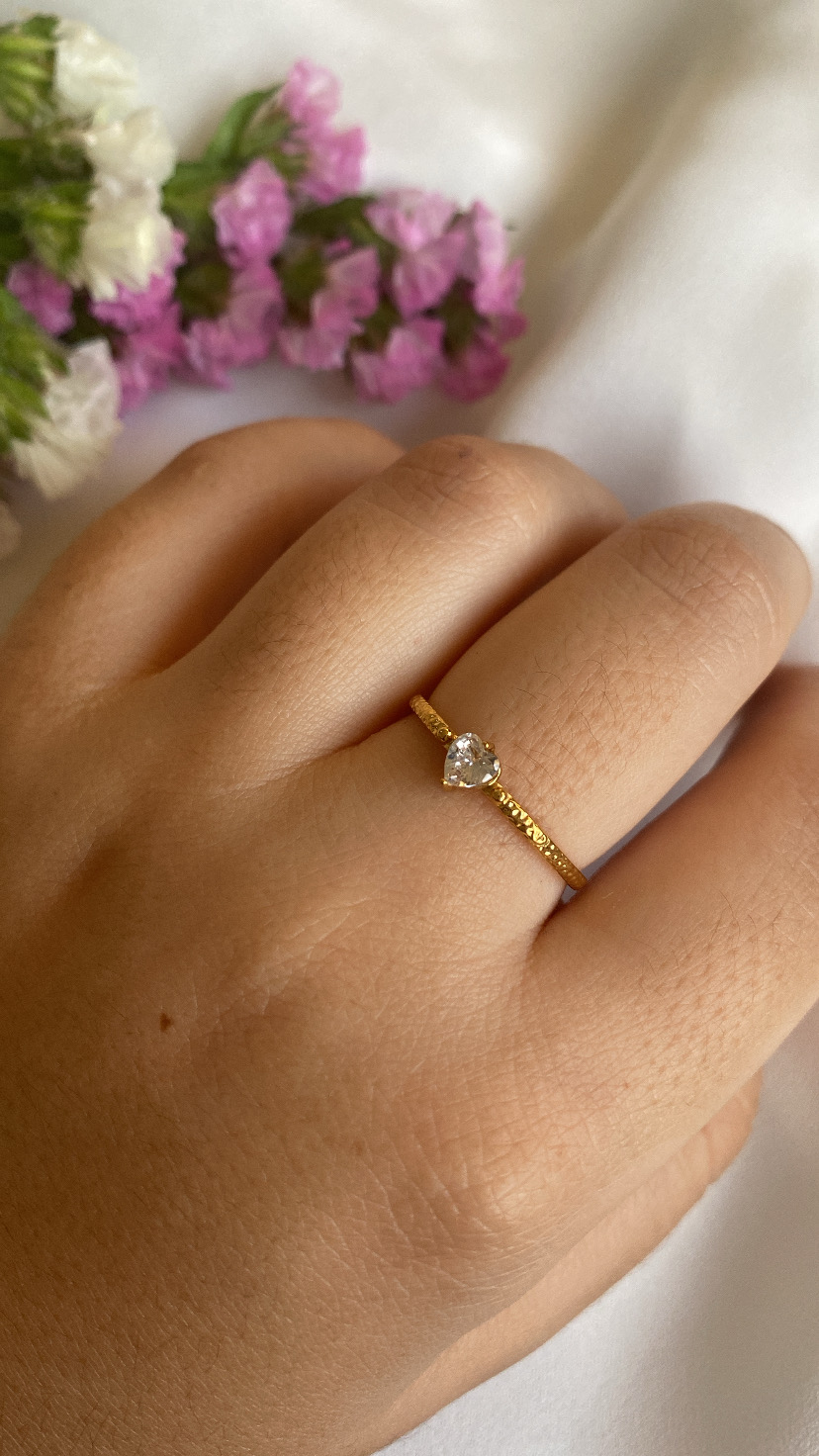 Anel dourado com pedra em forma de coração na mão, flores coloridas ao fundo