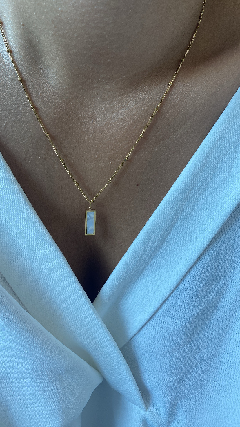 Colar dourado com pendente branco retangular num pescoço com camisa branca