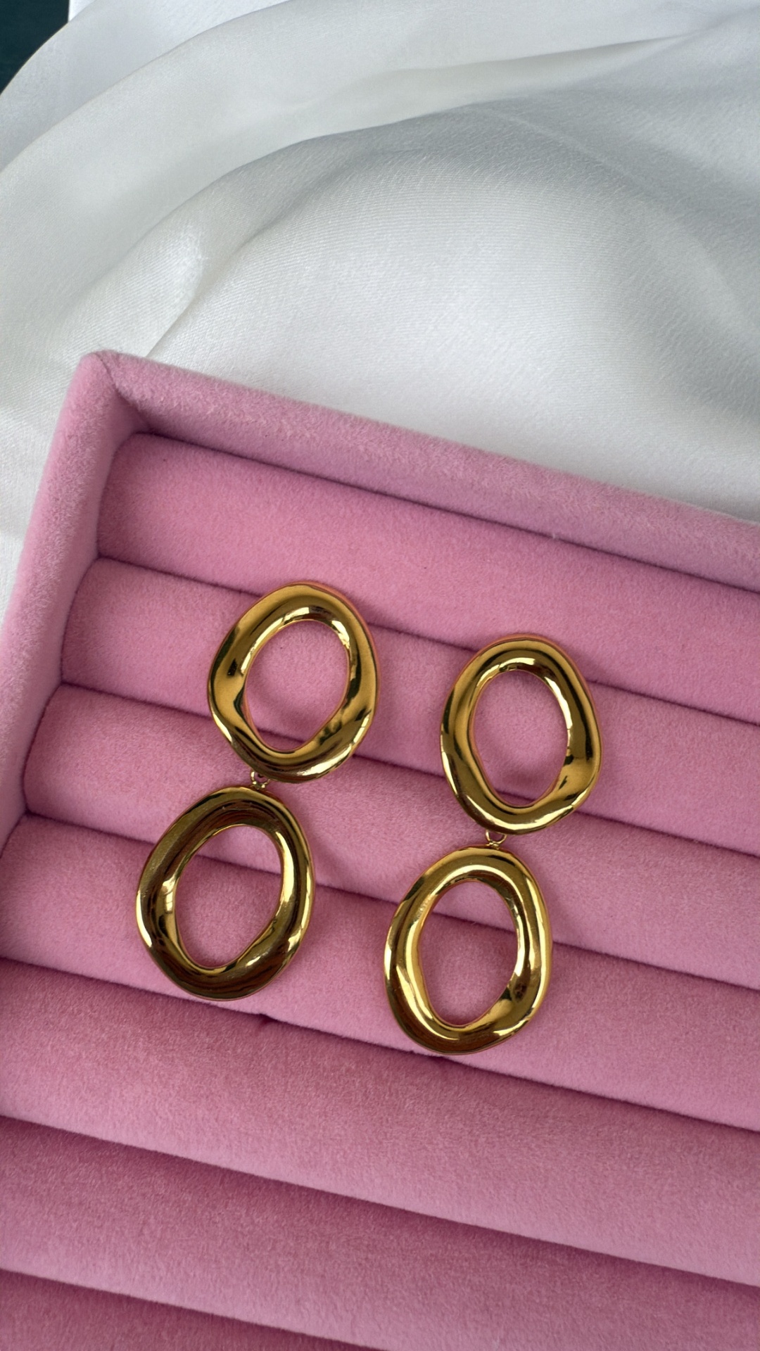 Brincos dourados ovalados sobre suporte rosa acolchoado