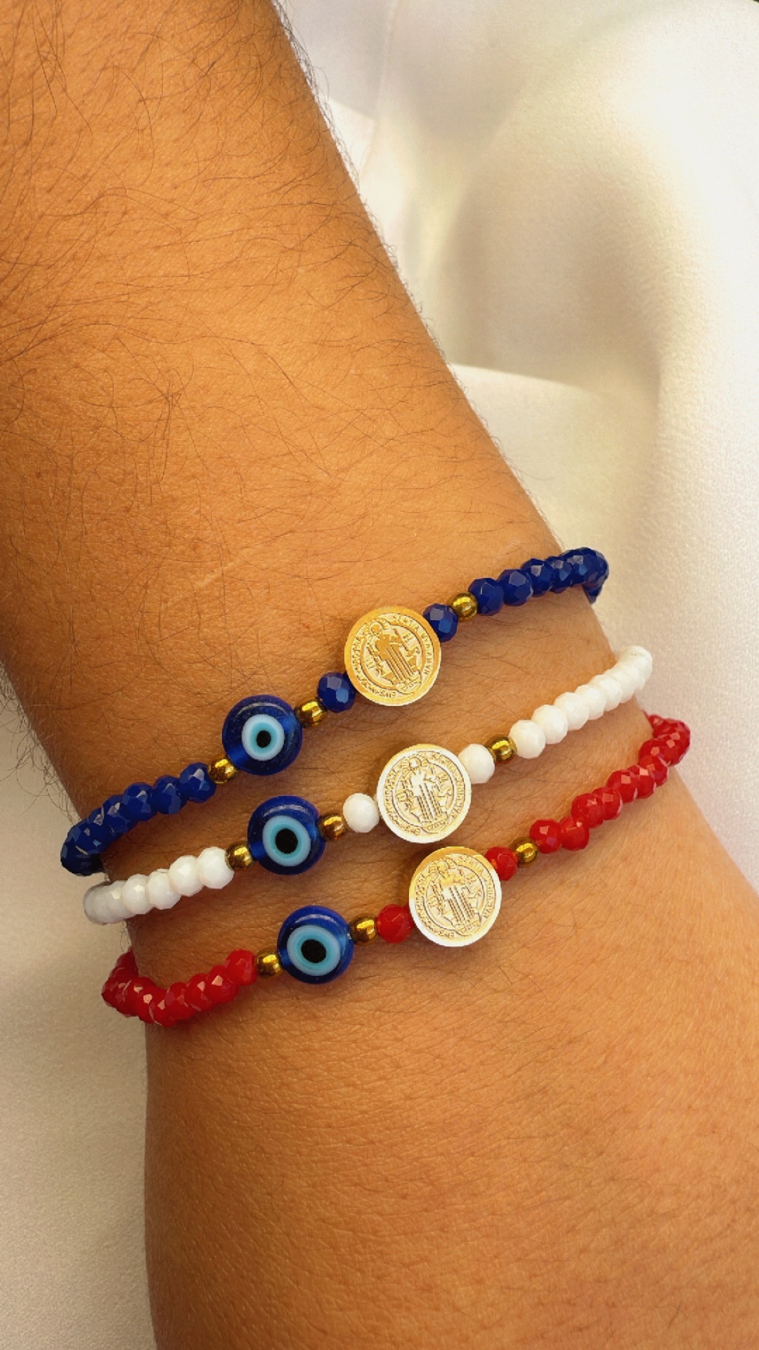 Três pulseiras coloridas com contas azuis, brancas e vermelhas, incluindo olhos gregos e moedas douradas, num pulso.