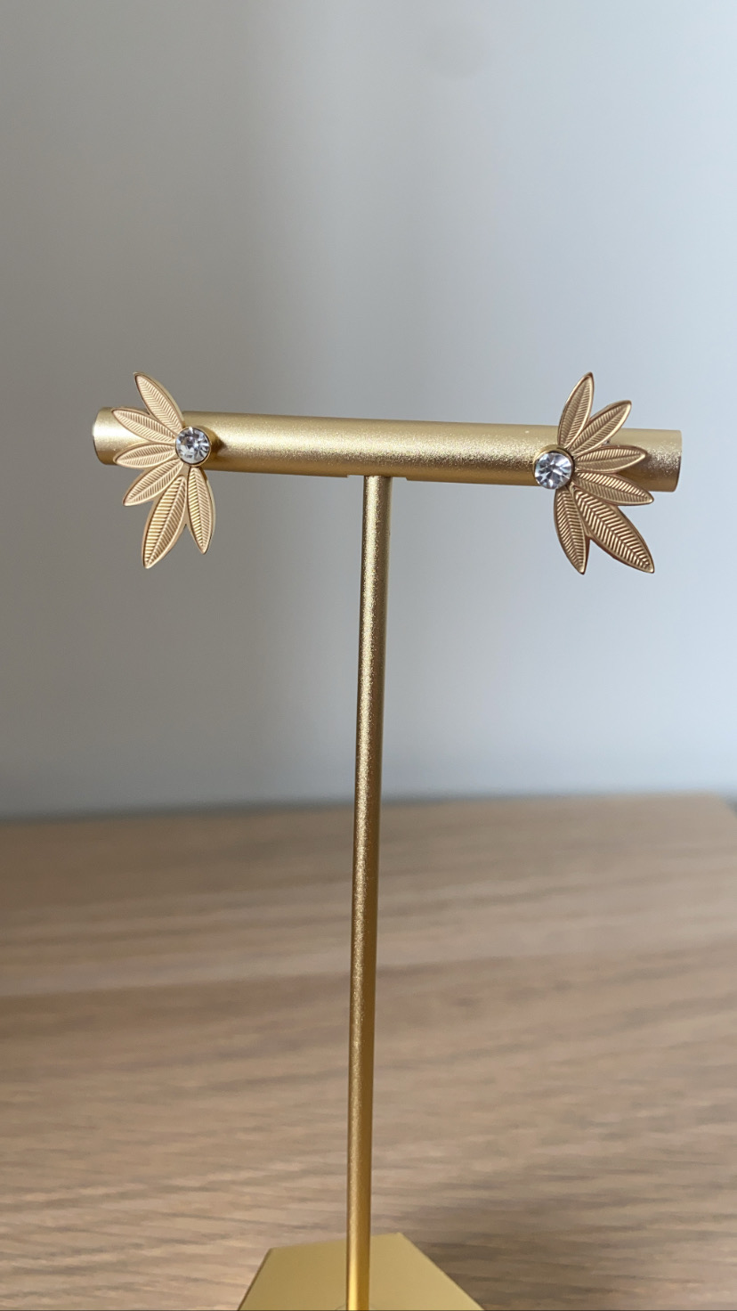 Brincos dourados em forma de folhas com pedras transparentes no centro, mostrados num suporte dourado