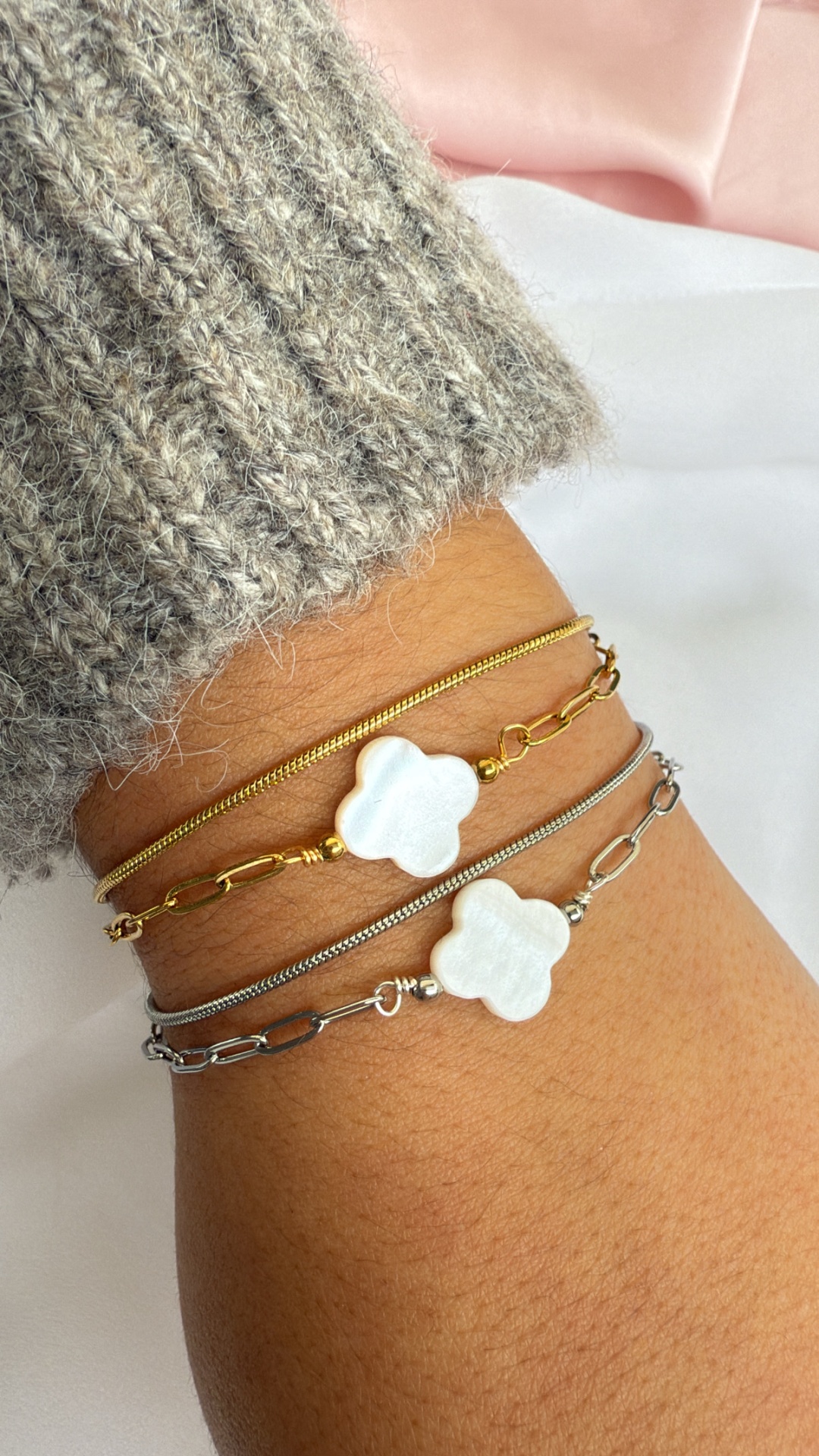 Pulseiras dourada e prateada com pendente de flor branca no pulso, com suéter cinza e fundo rosa e branco