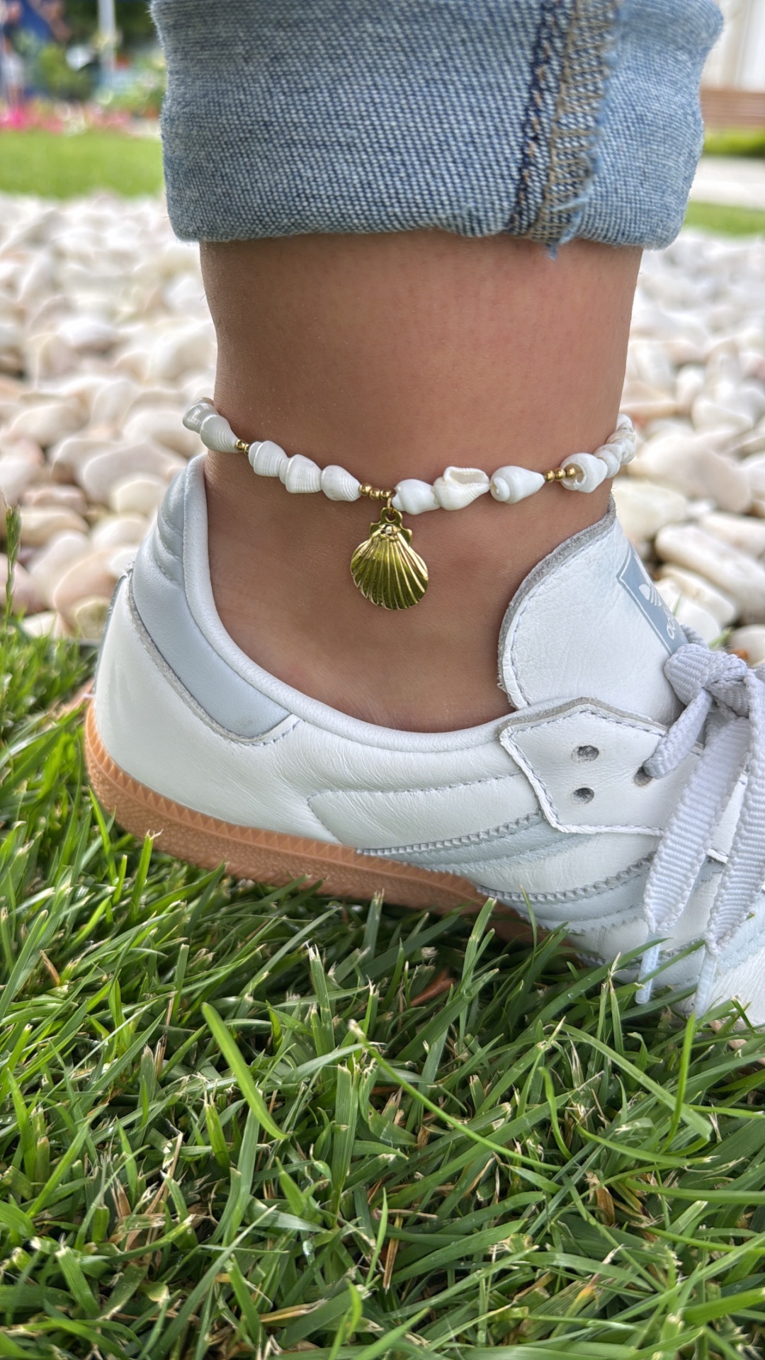 tornozeleira com pedras brancas e pendente de concha dourada junto a sapatilha branca no relvado