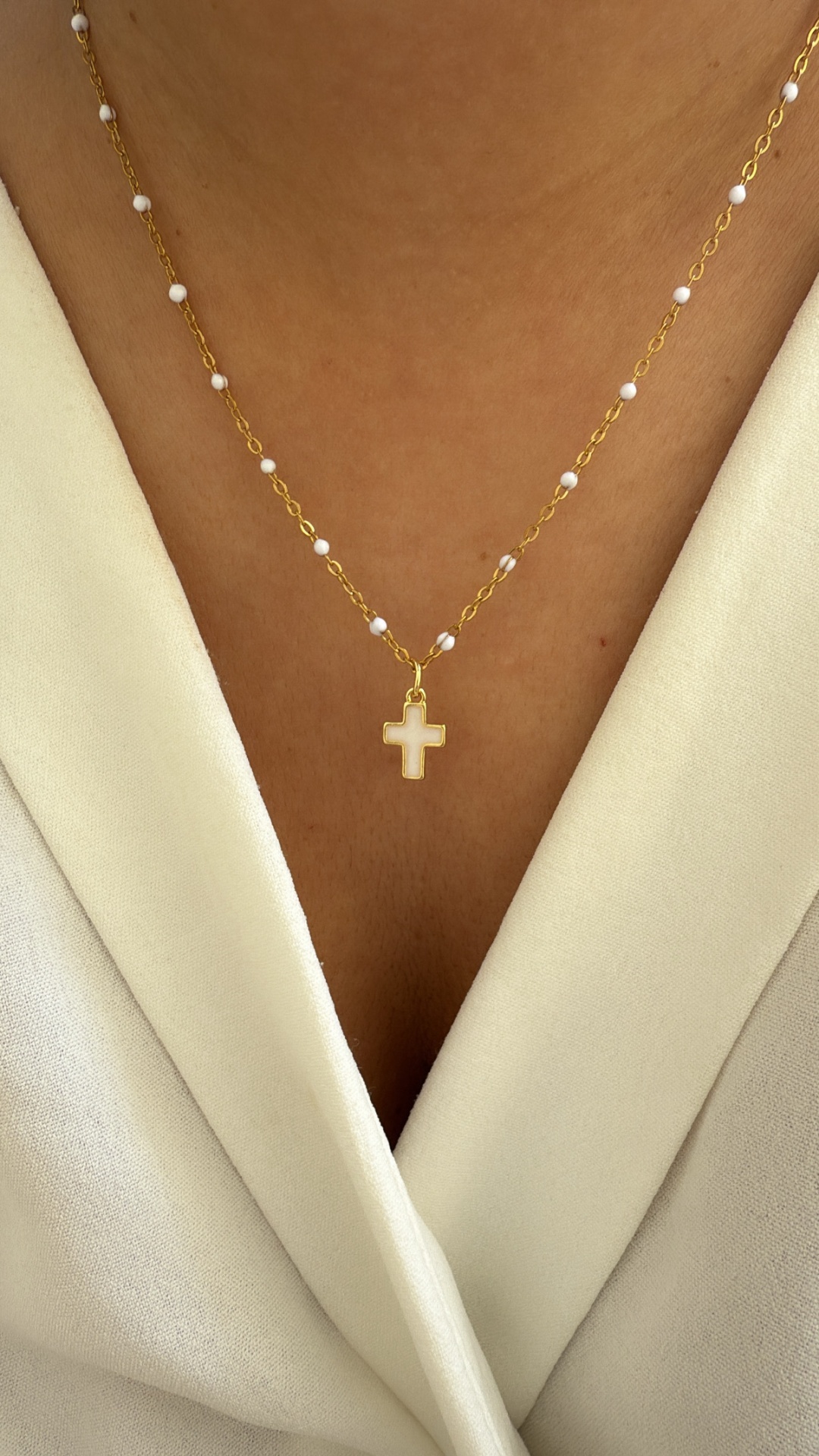 Colar dourado com contas brancas e pendente em cruz creme sobre pele e vestido branco