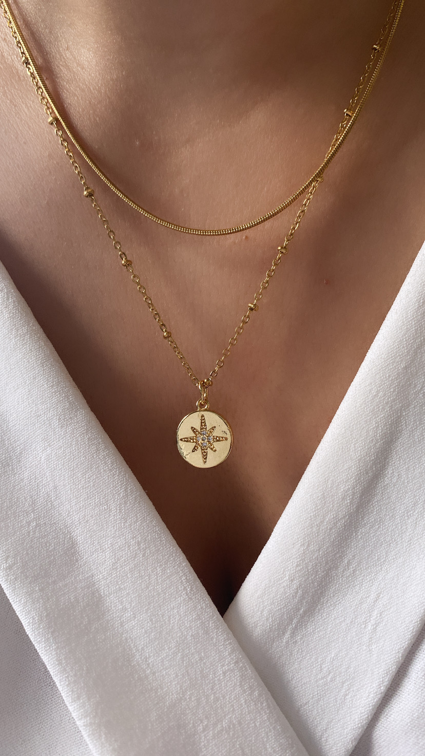 Colar dourado com pendente redondo e estrela de oito pontas com pedras sobre tecido branco