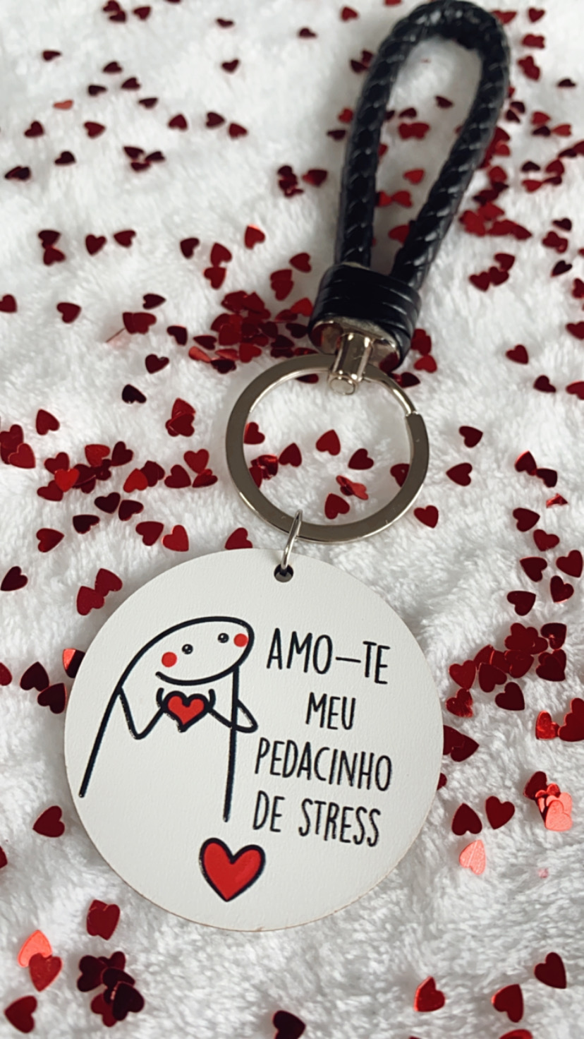 Chaveiro redondo branco com figura e texto, argola metálica e cordão preto em fundo com corações vermelhos
