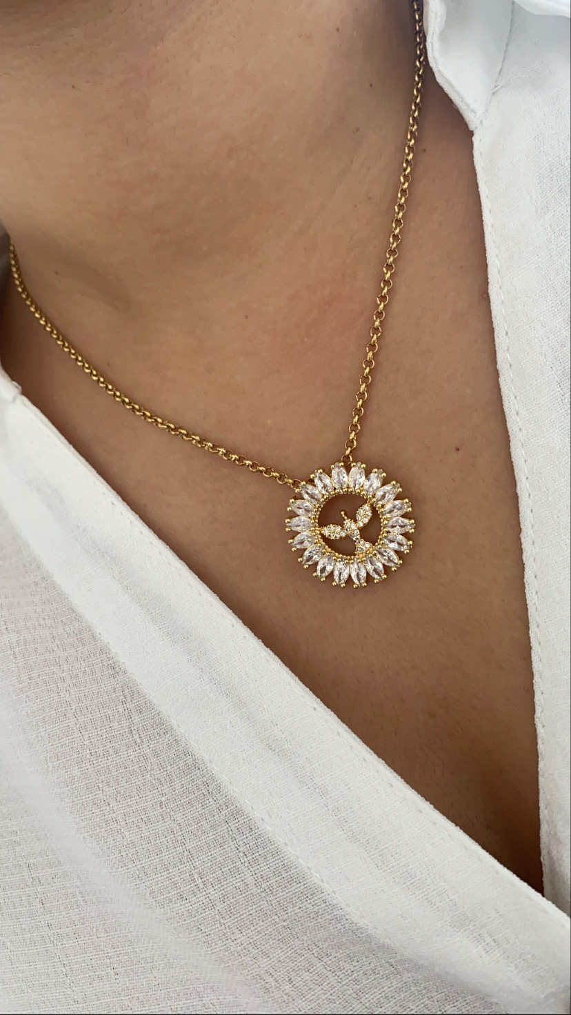 Colar dourado com pendente circular de pedras brancas sobre roupa branca