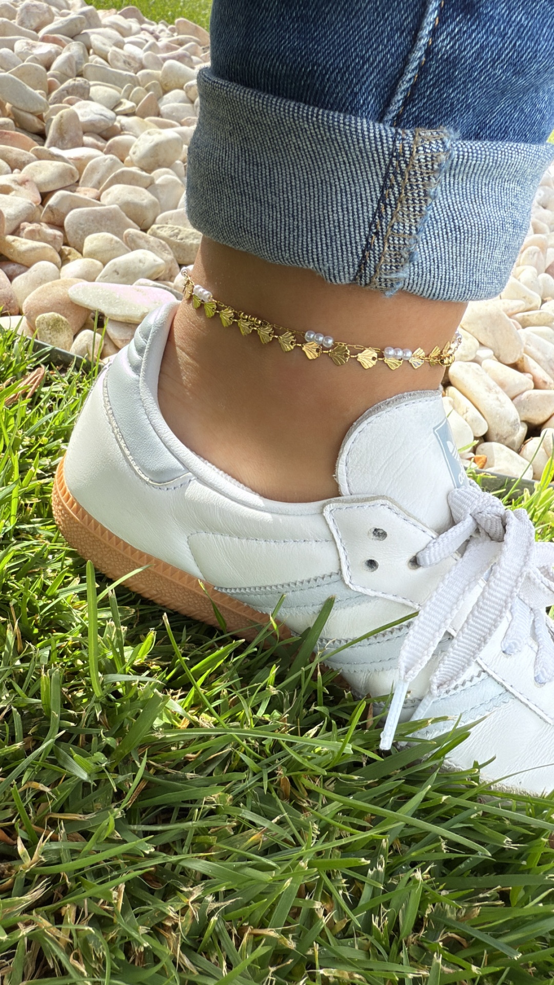 Ténis brancos em pele com sola castanha e tornozeleira dourada na perna sobre relva e pedras brancas