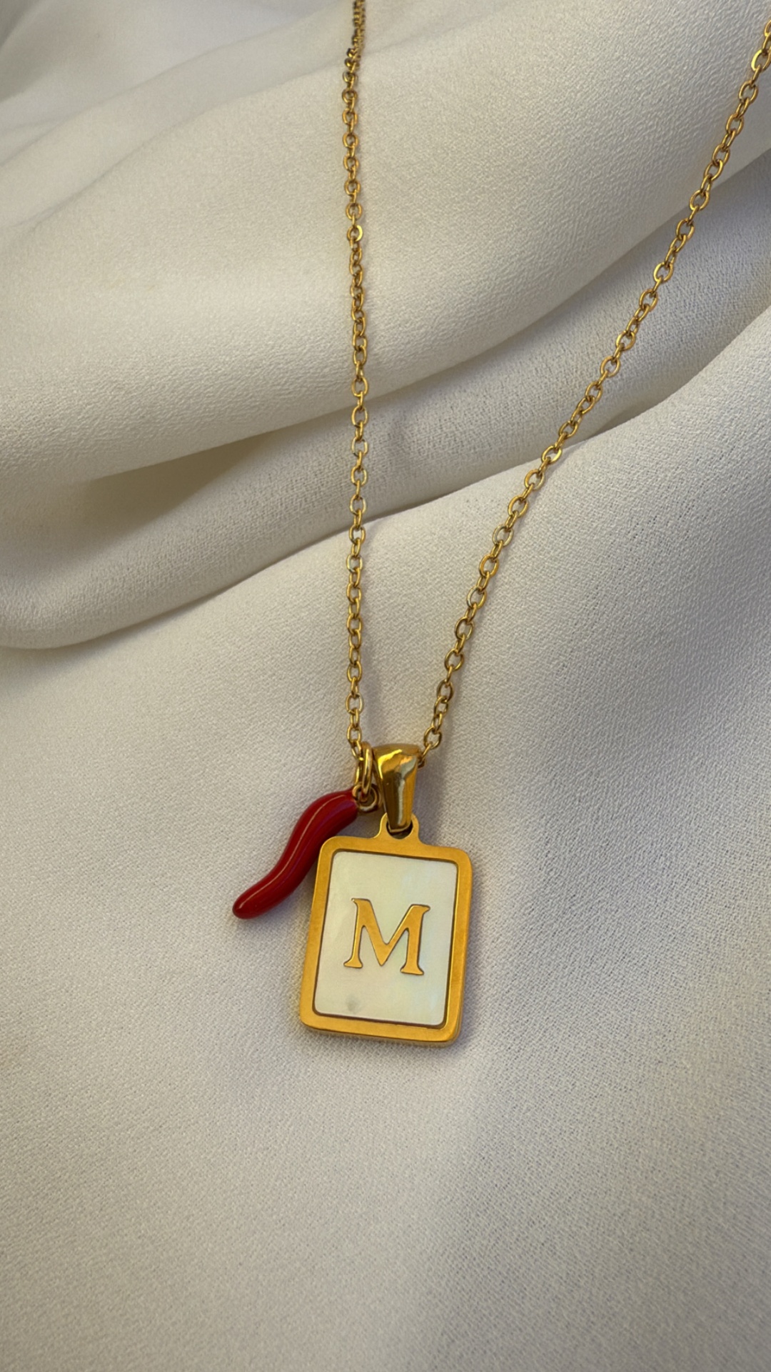 Colar dourado com pendente retangular branco com a letra M dourada e pendente vermelho em forma de pimento