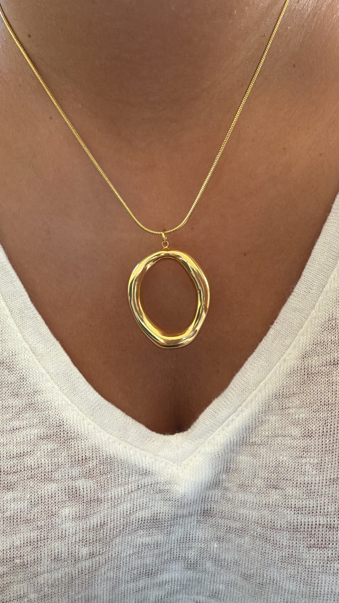 Colar dourado com pendente oval irregular e camisola branca de malha.