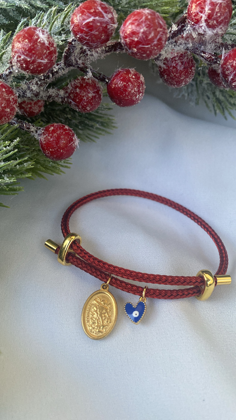 Pulseira vermelha com medalhão dourado e coração azul pendurados perto de ramos de pinheiro e bagas vermelhas artificiais