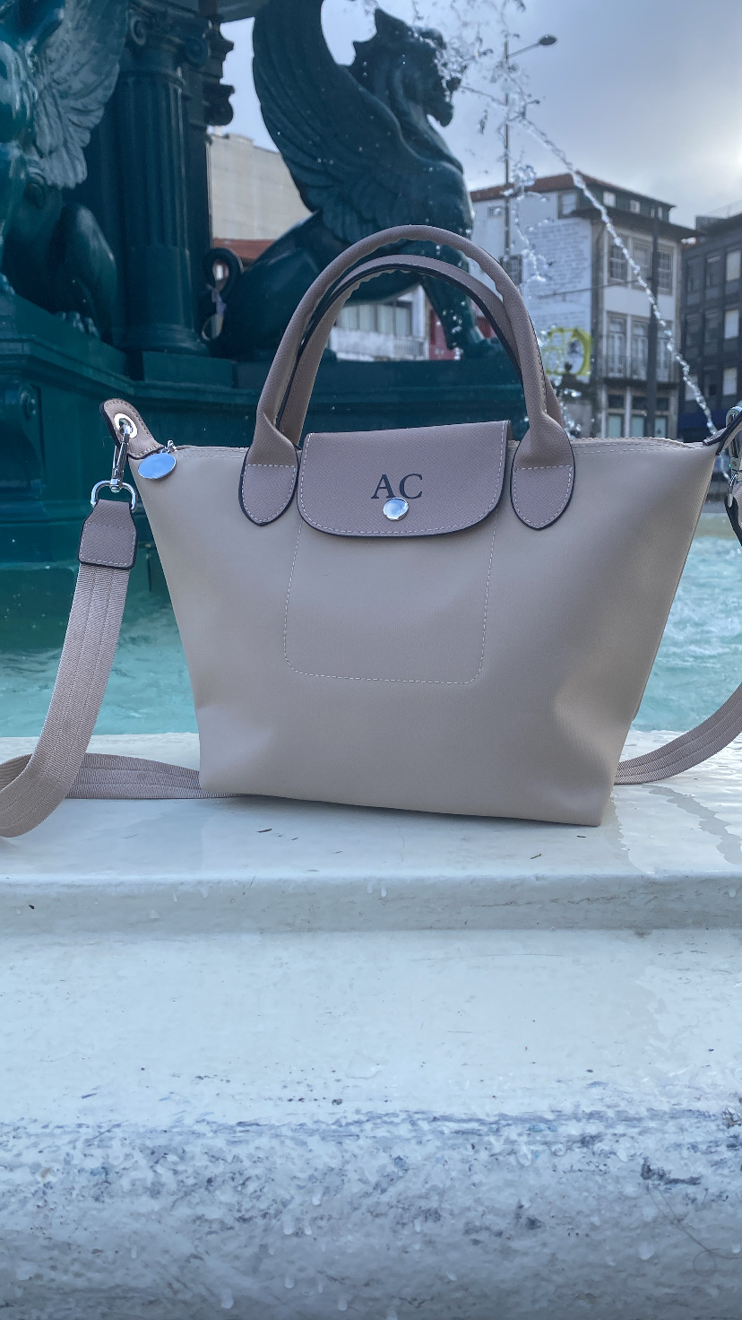 Mala de mão bege com alças e correia castanha junto a fonte com estátuas negras de cavalos alados