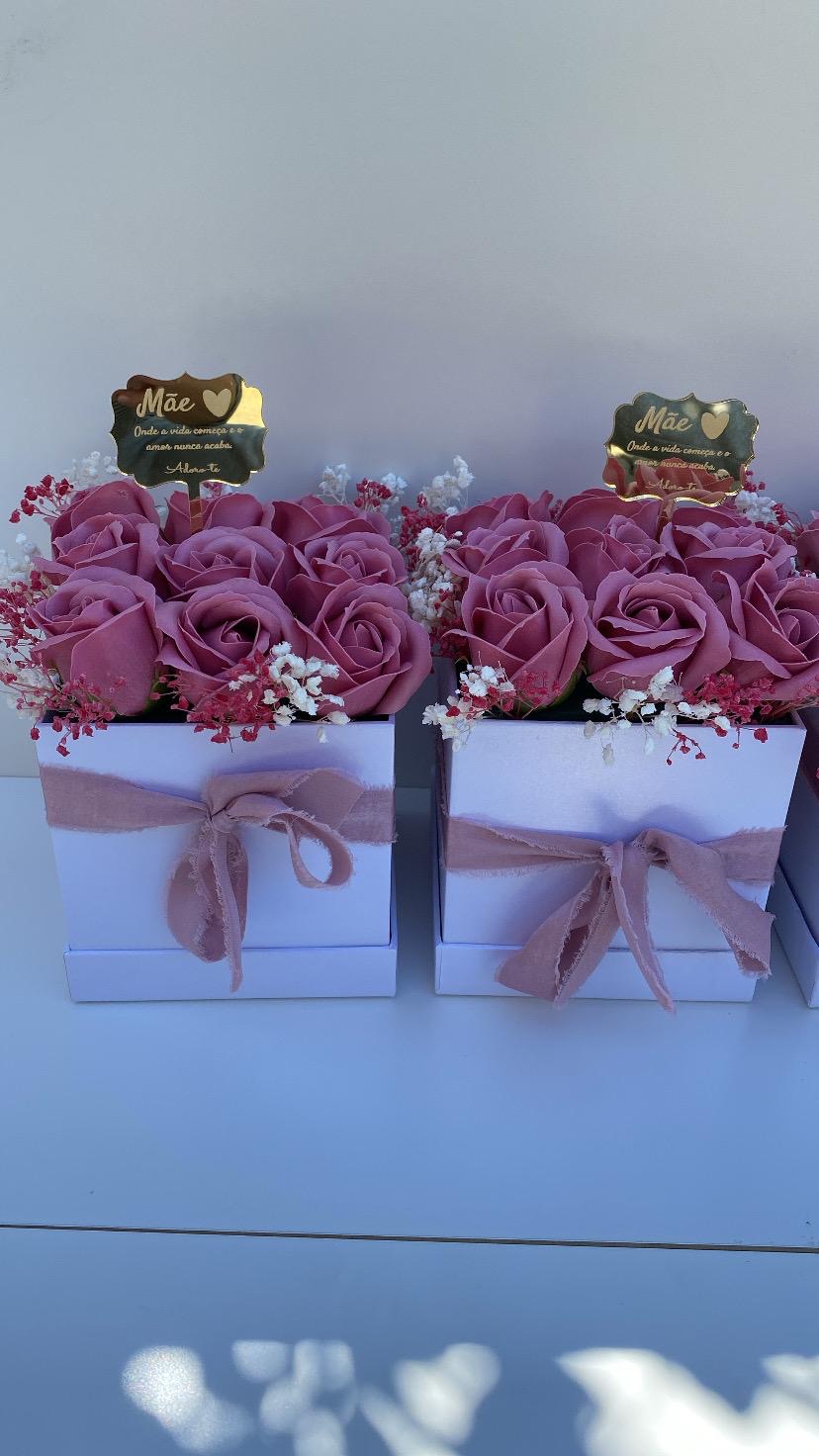Dois arranjos florais em caixas brancas com laços cor-de-rosa e uma placa dourada com texto para mãe.