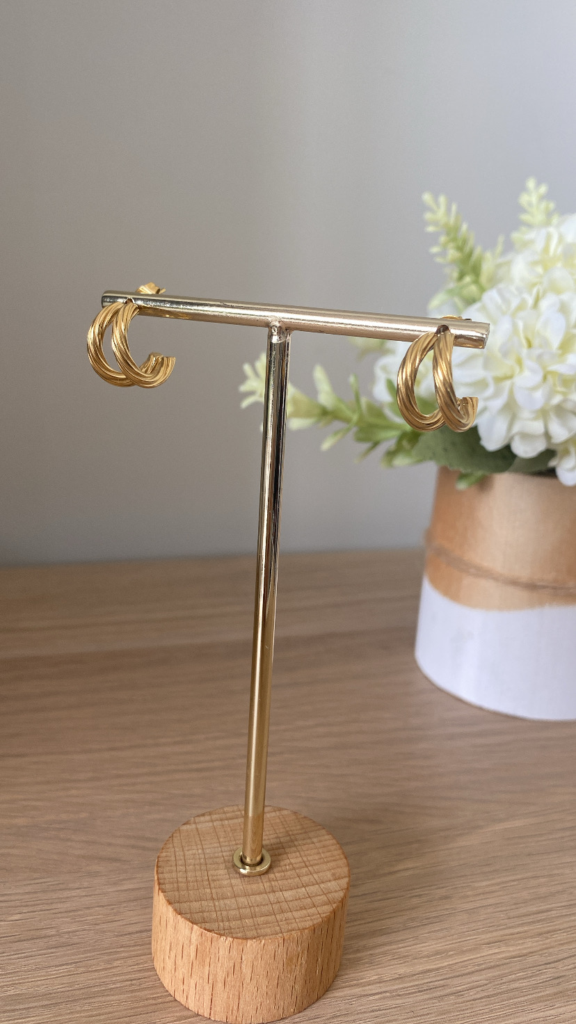 Brincos dourados em suporte metálico com base de madeira e planta ao fundo