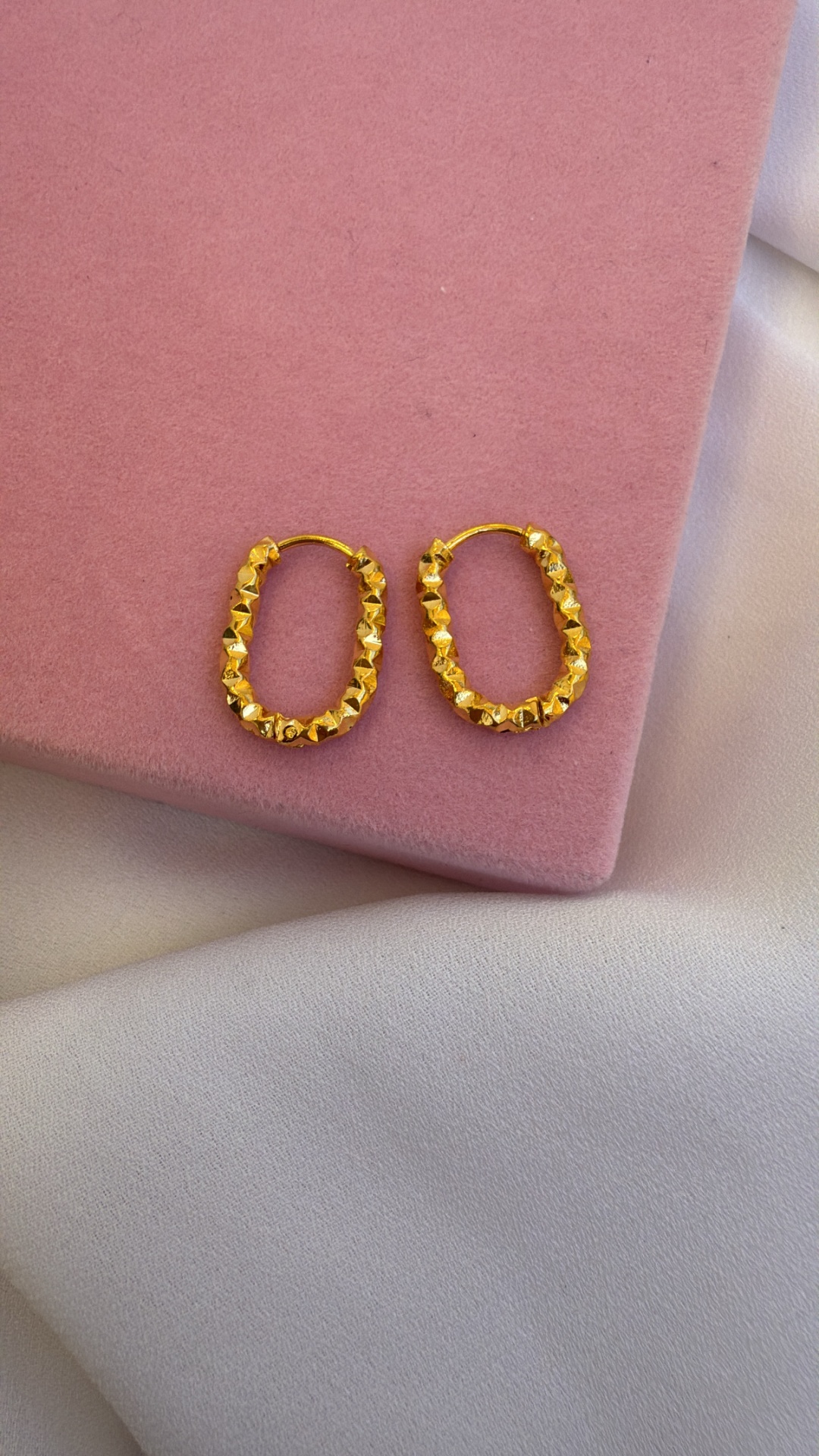 Brincos dourados ovalados com textura facetada sobre superfície rosa aveludada