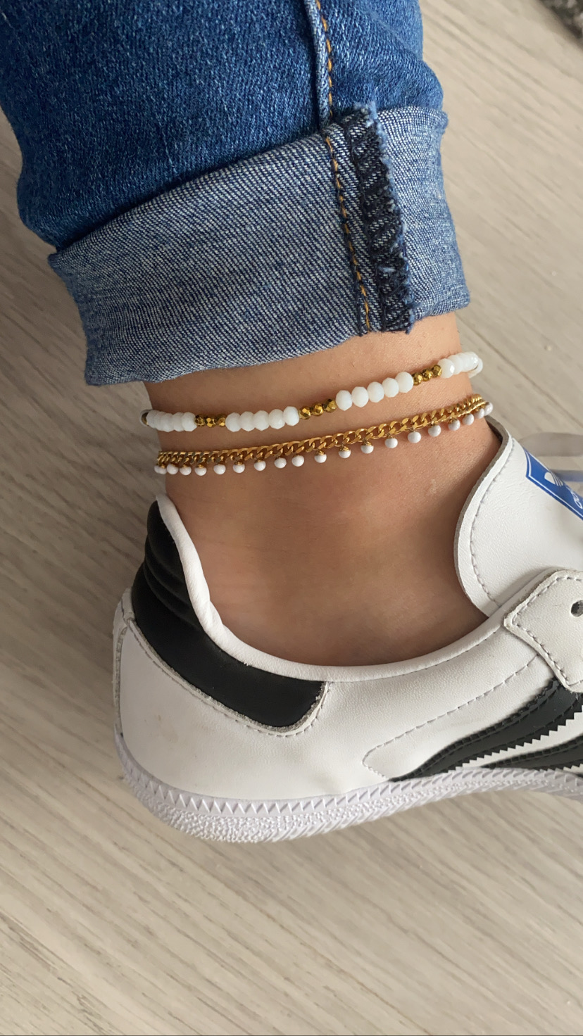 Tornozelo com pulseiras douradas e brancas, jeans azul e ténis branco com detalhes pretos