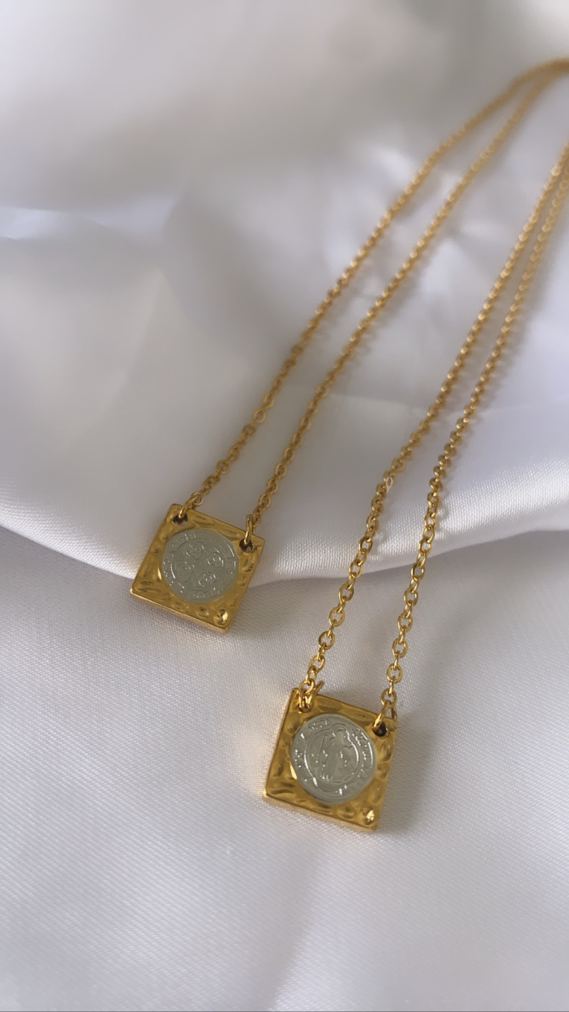 Duas colares dourados com pendentes quadrados e moedas prateadas no centro sobre tecido branco