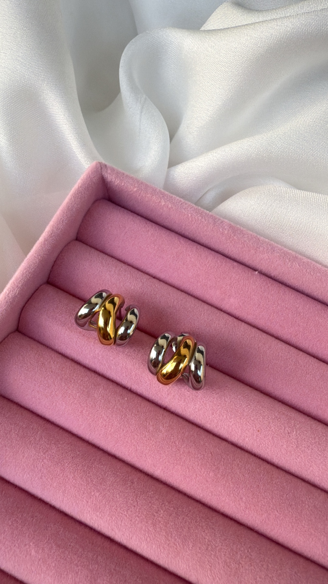 Brincos bicolores de metal numa caixa rosa de veludo sobre fundo branco.