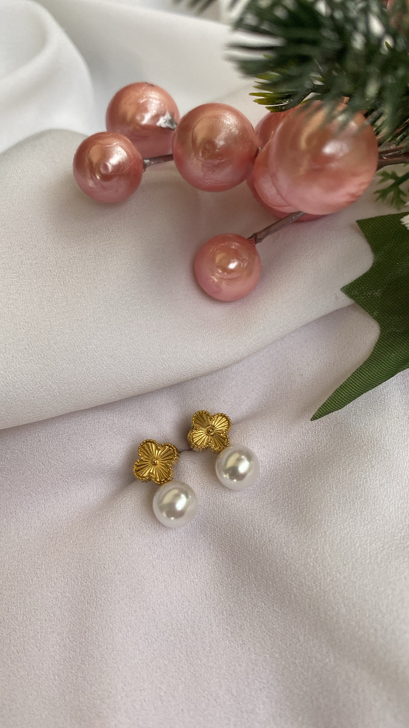 Brincos dourados em forma de flor com pérolas brancas, sobre tecido branco com decoração floral rosa e verde