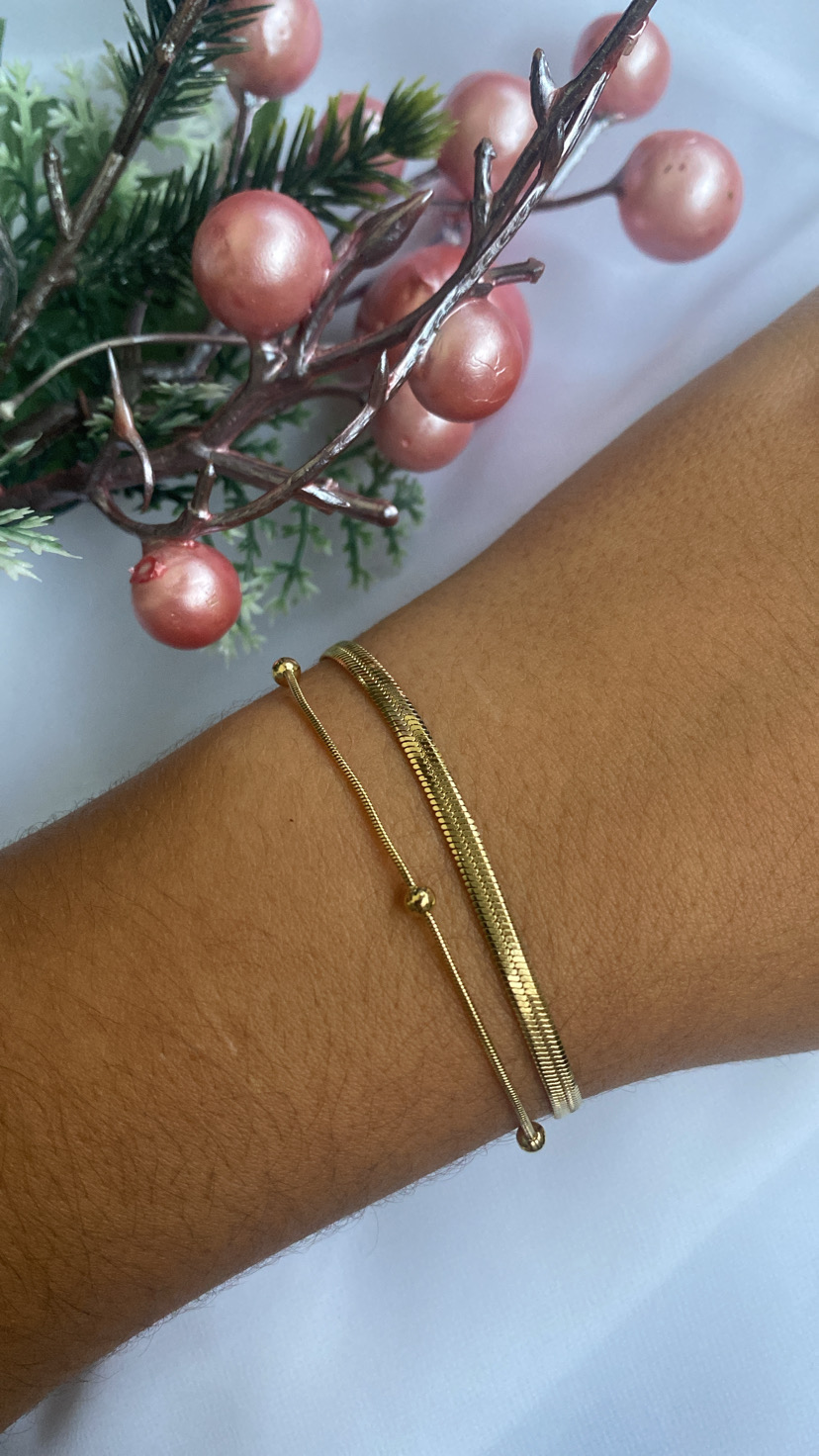 Pulso com duas pulseiras douradas perto de ramos decorativos rosa e verdes