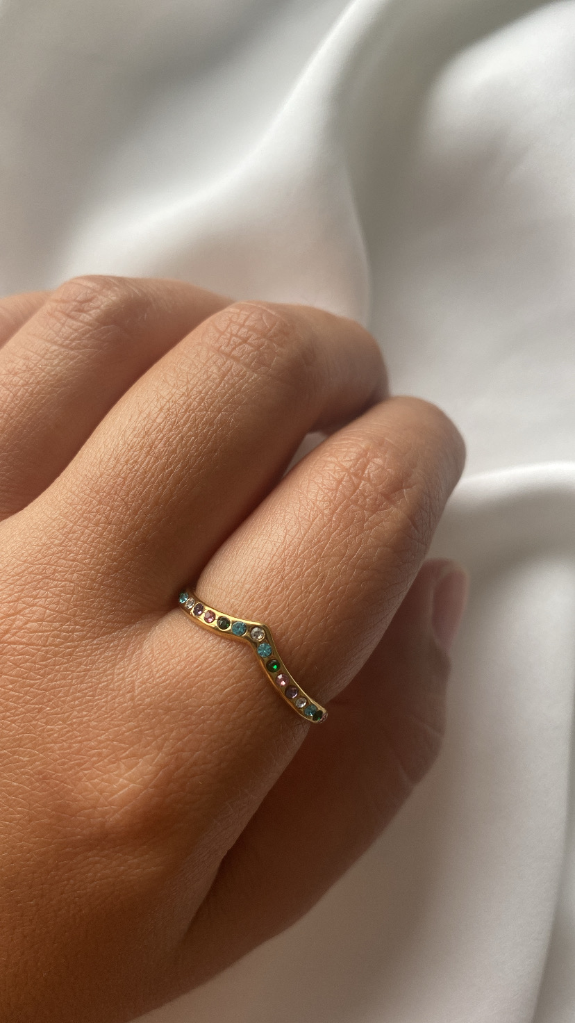 Anel dourado com pedras coloridas em formato de V no dedo médio, sobre tecido branco