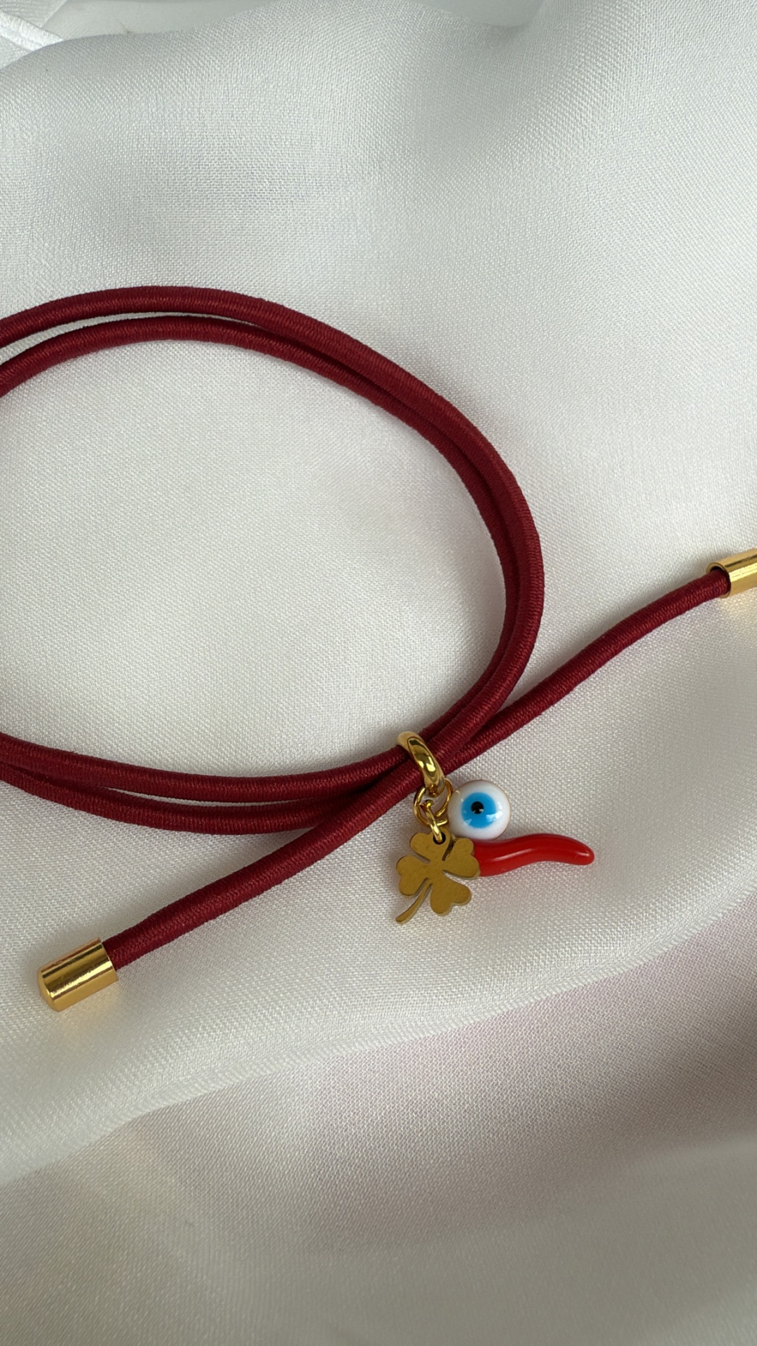 Pulseira vermelha com pendentes dourado, olho azul e chifre vermelho sobre tecido branco