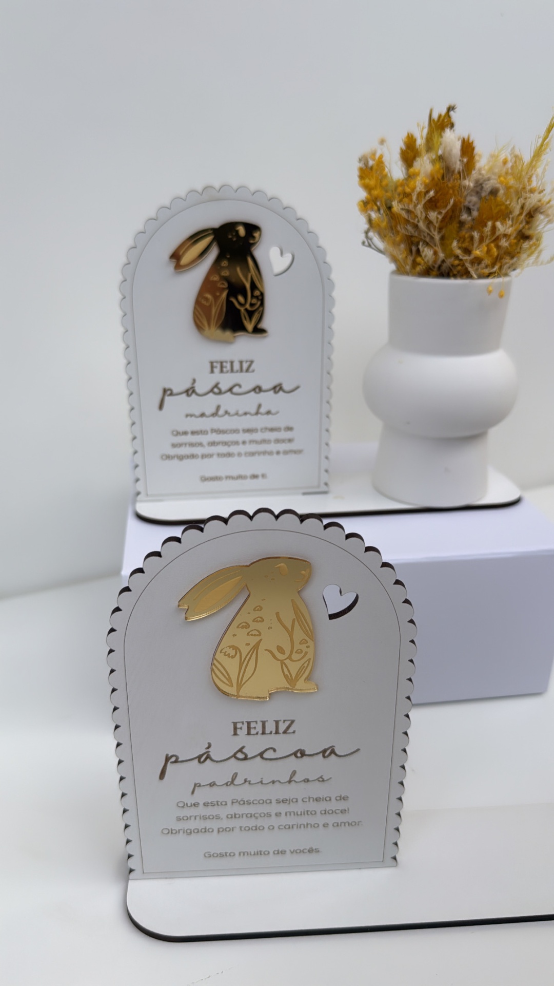 Placas decorativas de Páscoa com coelhos dourados e texto em português em fundo branco com vaso e flores secas ao fundo
