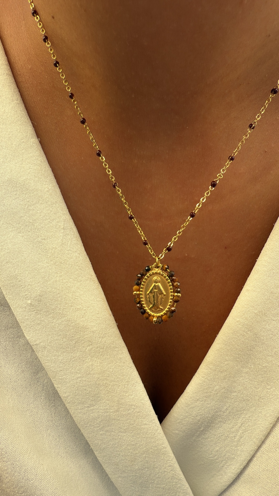 Colar dourado com pendente oval religioso e pequenas contas castanhas, usado com casaco claro.