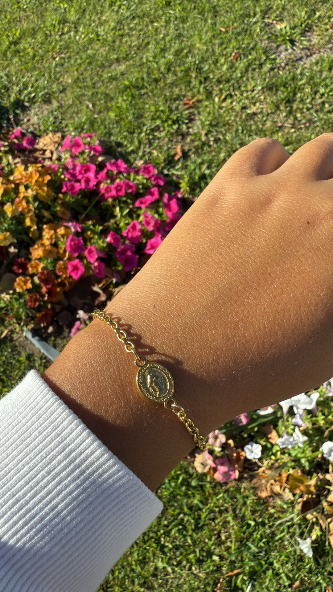 Pulseira dourada fina com pendente oval gravado num pulso com fundo de flores e relva