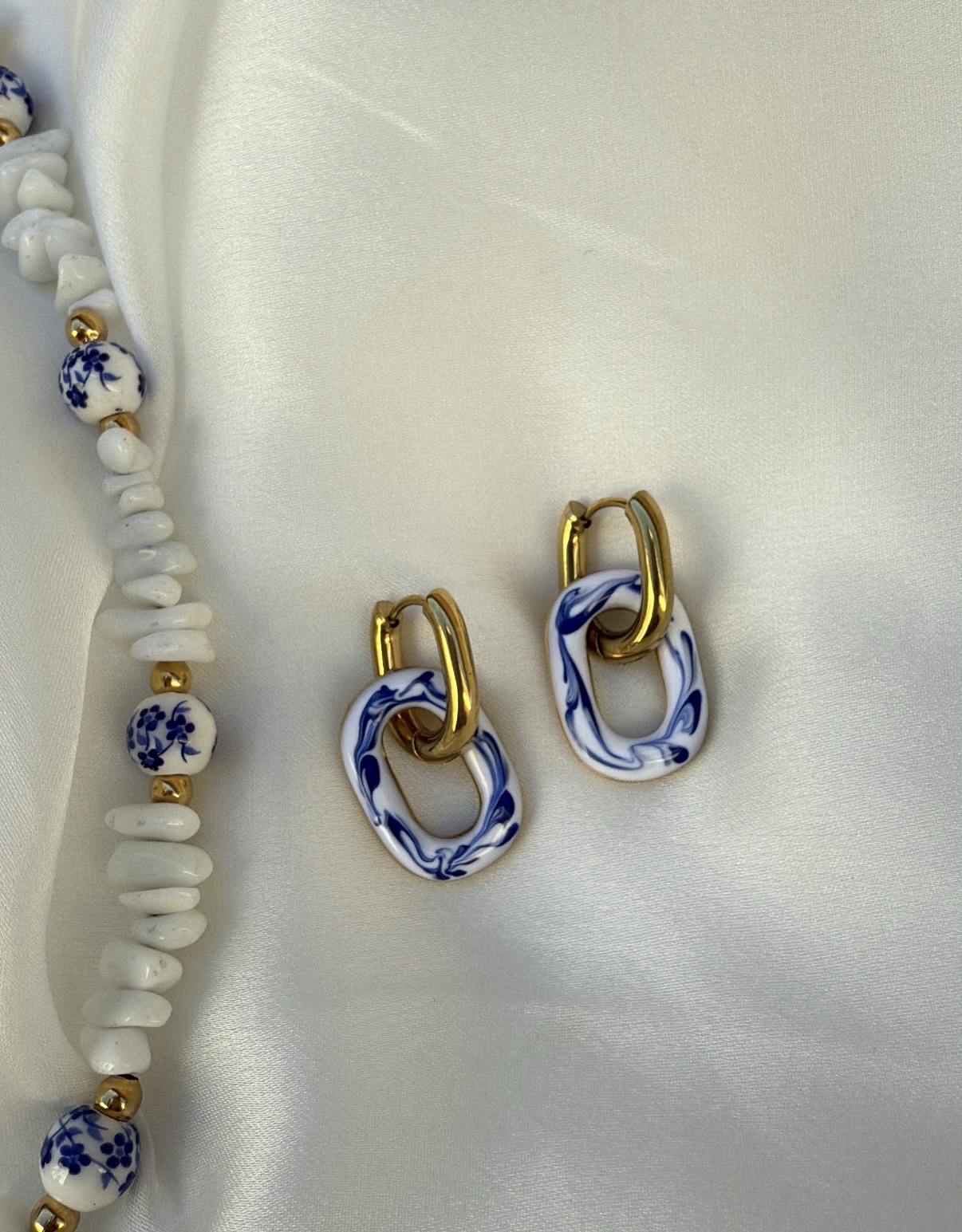 Brincos dourados com detalhes azul e branco e colar de contas brancas e douradas sobre tecido branco