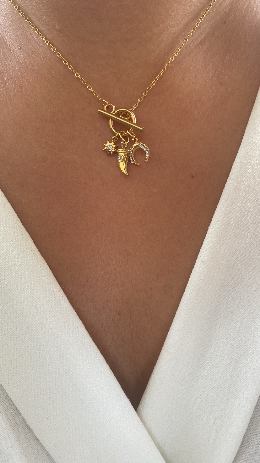 Colar dourado com pendentes de estrela, dente e ferradura no pescoço de pessoa de pele morena e roupa branca.