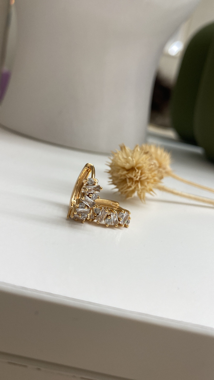 Brinco dourado com pedras transparentes sobre superfície branca com plantas secas ao fundo