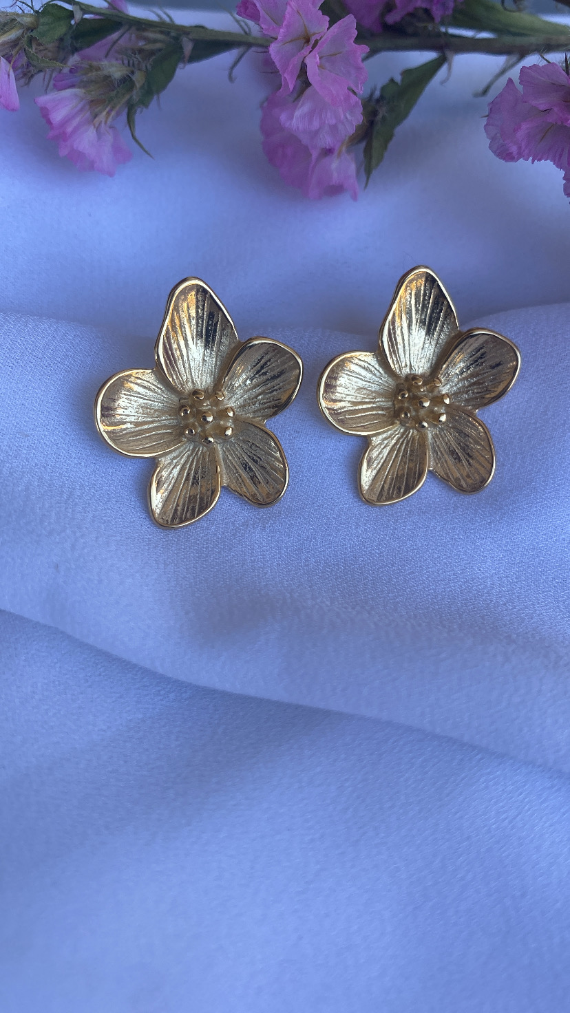 Brincos flor dourados sobre tecido branco com flores rosas ao fundo