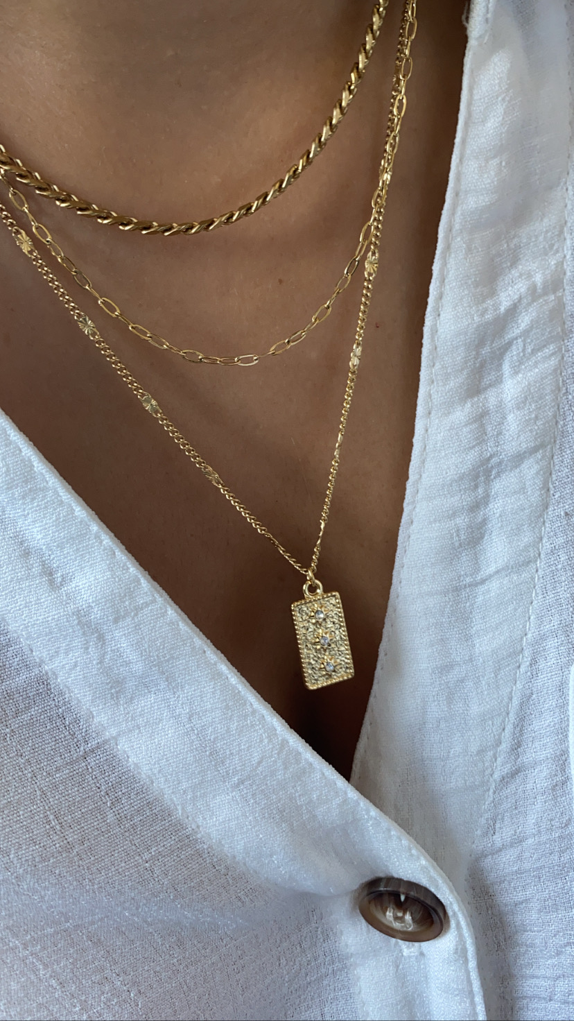 Colar dourado em camadas com pendente retangular brilhante, usado sobre roupa branca com botão.