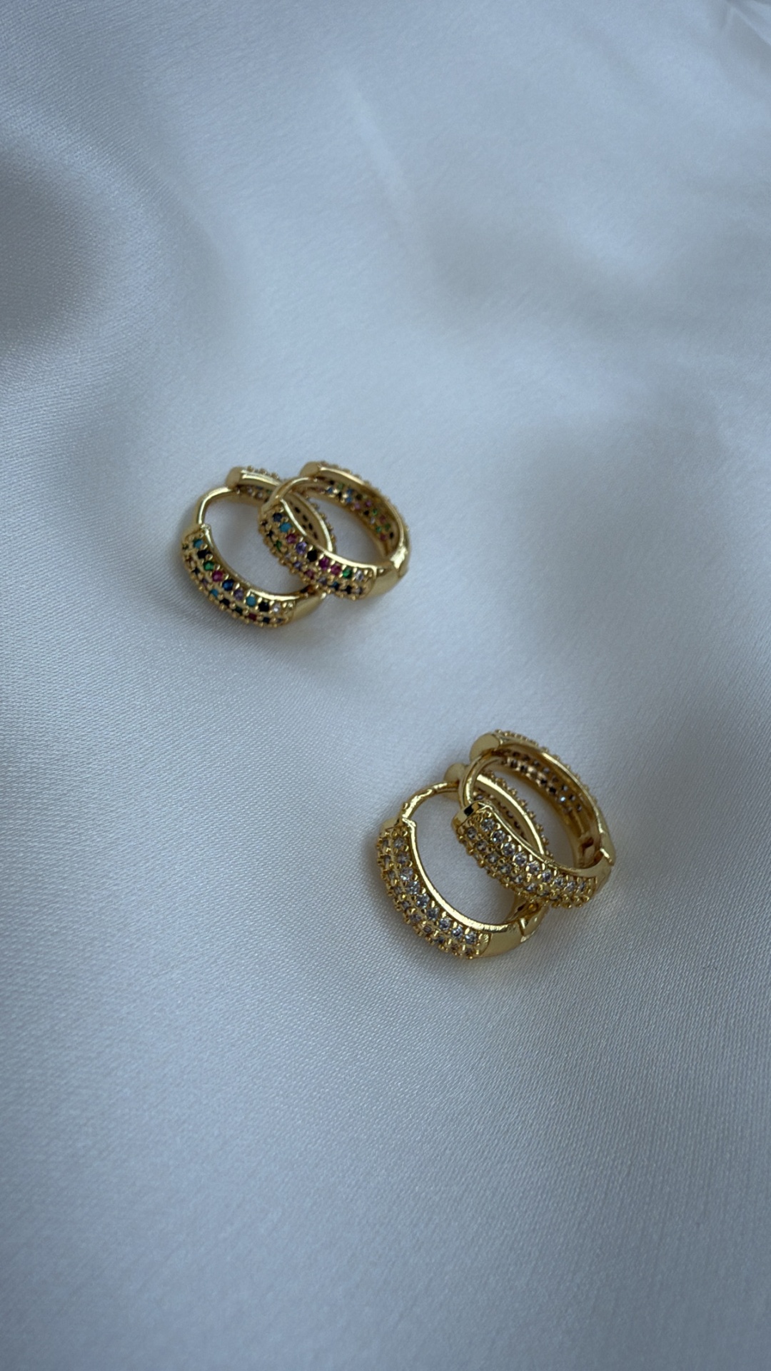 Dois pares de brincos argola dourados com pedras coloridas e brancas sobre tecido branco.