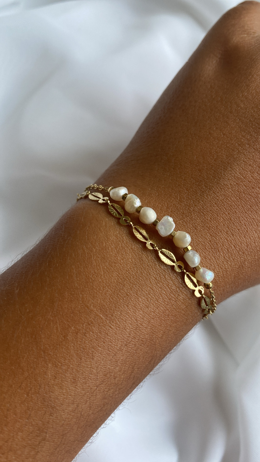 Bracelete duplo dourado com pérolas num pulso sobre fundo branco