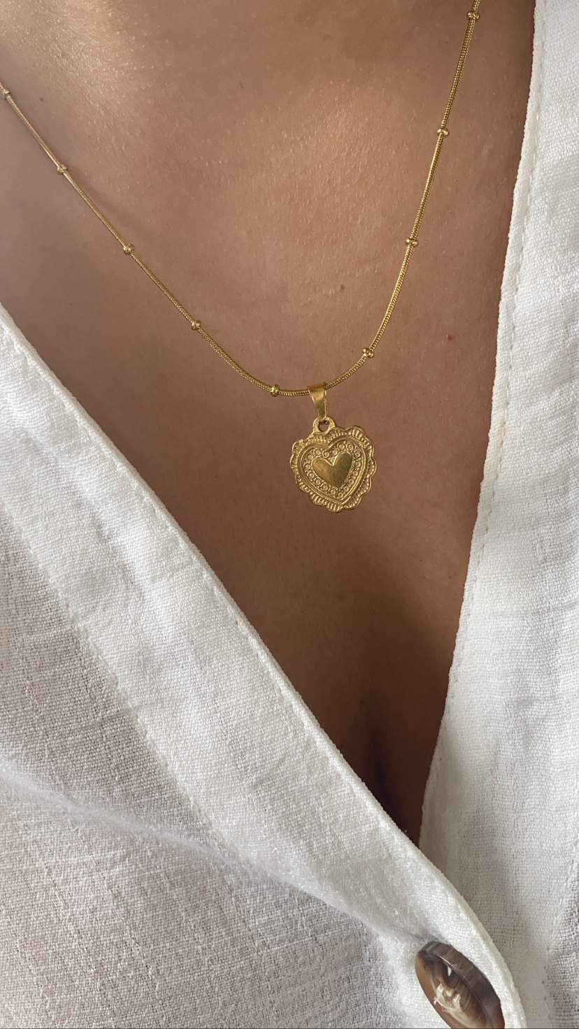 Colar dourado com pendente de coração em corrente fina sobre pele e camisa branca