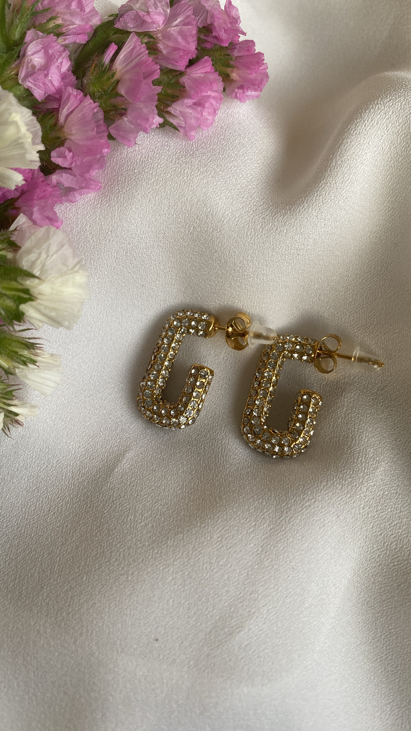 Brincos dourados em forma de letra G com cristais sobre tecido branco e flores secas cor-de-rosa e creme