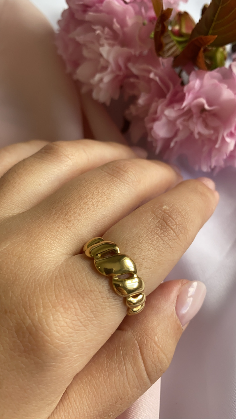 anel dourado ondulado em dedo com flores cor-de-rosa ao fundo
