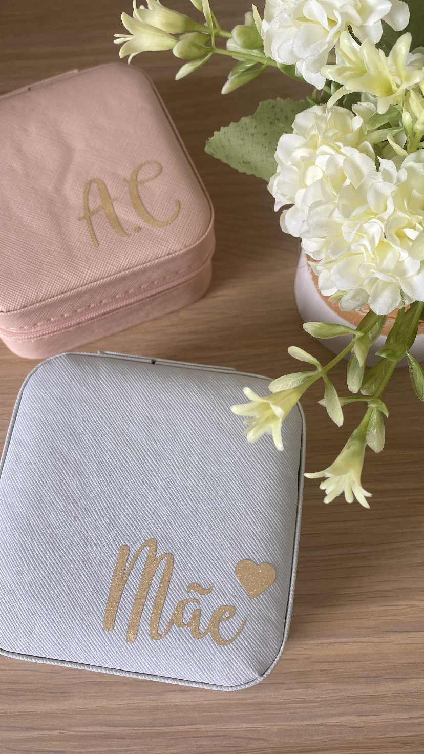 Duas bolsas quadradas pequenas, uma rosa com letras douradas AE e outra cinza com palavra Mãe e coração dourados, ao lado de flores brancas