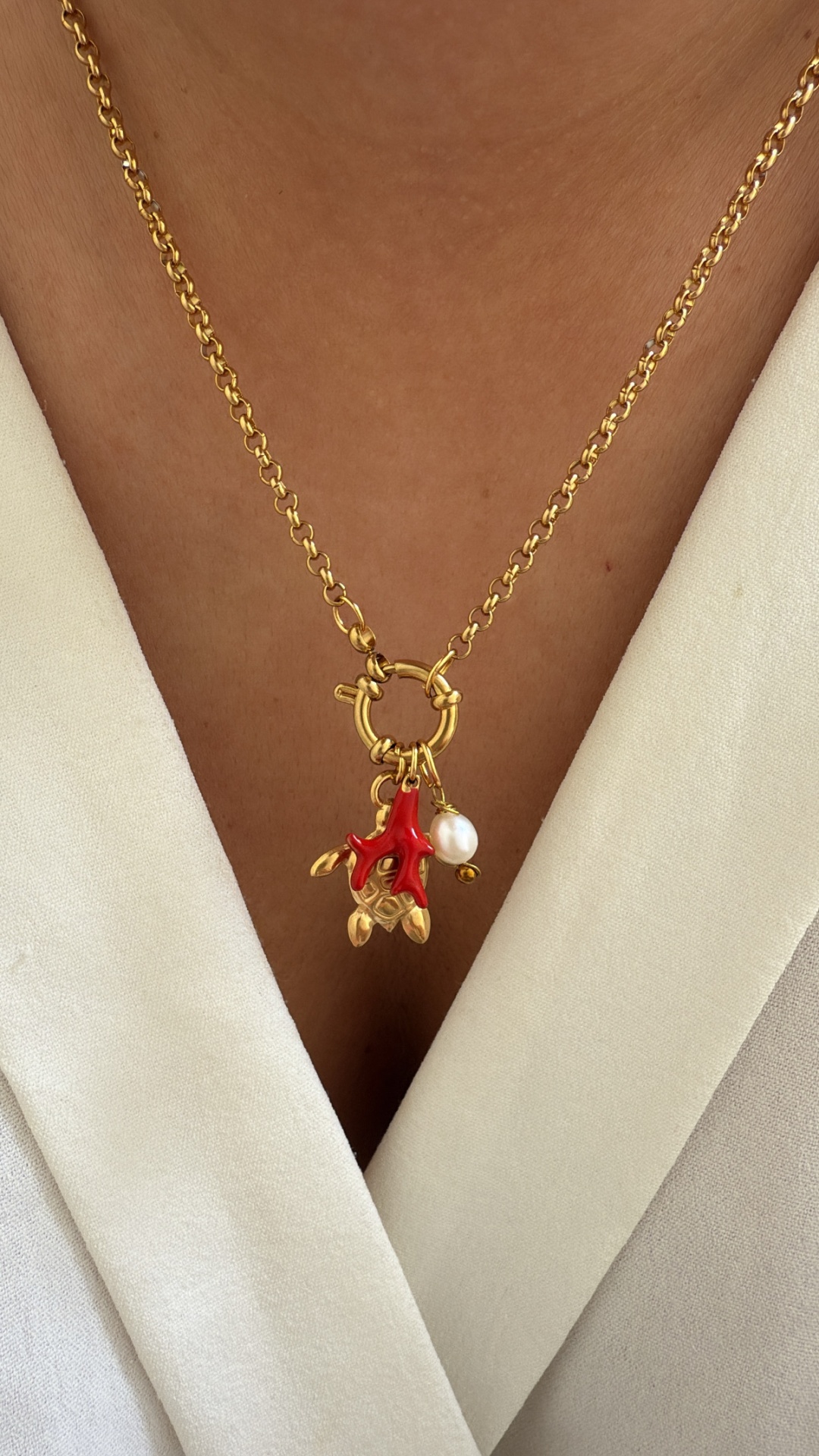 Colar dourado com pendente vermelho e pérola numa pessoa com camisa bege clara