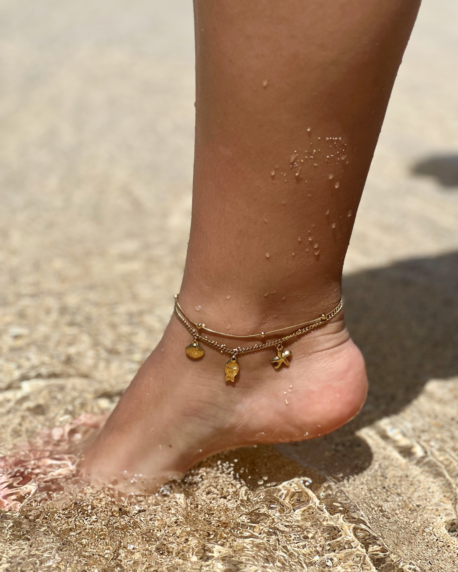 Tornozelo com pulseira dourada e pingentes na areia e água