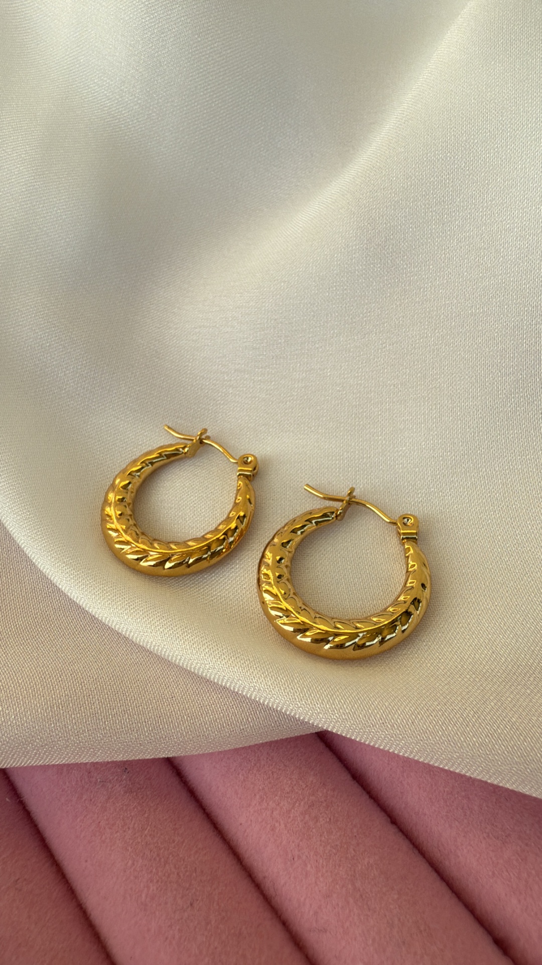 Brincos de argola dourados texturizados sobre tecidos branco e rosa