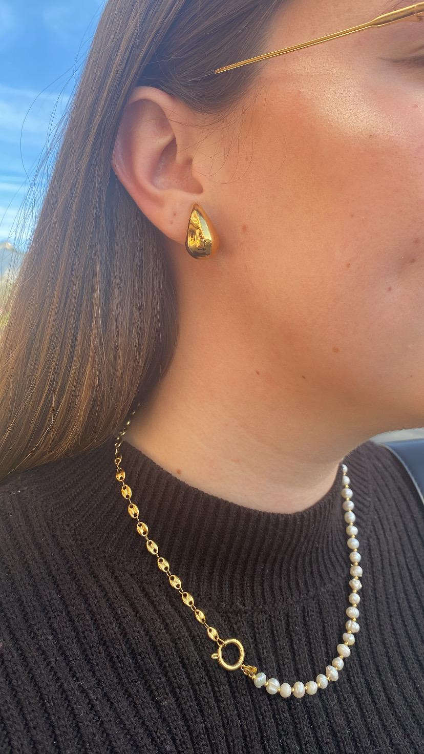 Brincos dourados irregulares e colar de pérolas com corrente dourada em mulher com camisola preta