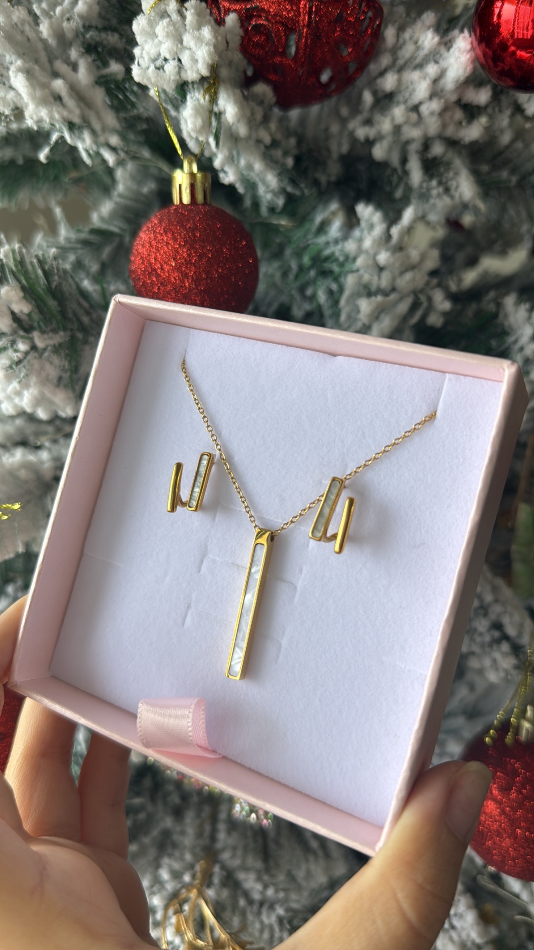 Conjunto de colar e brincos dourados com detalhes brancos numa caixa rosa, frente a árvore de Natal nevado com bolas vermelhas.