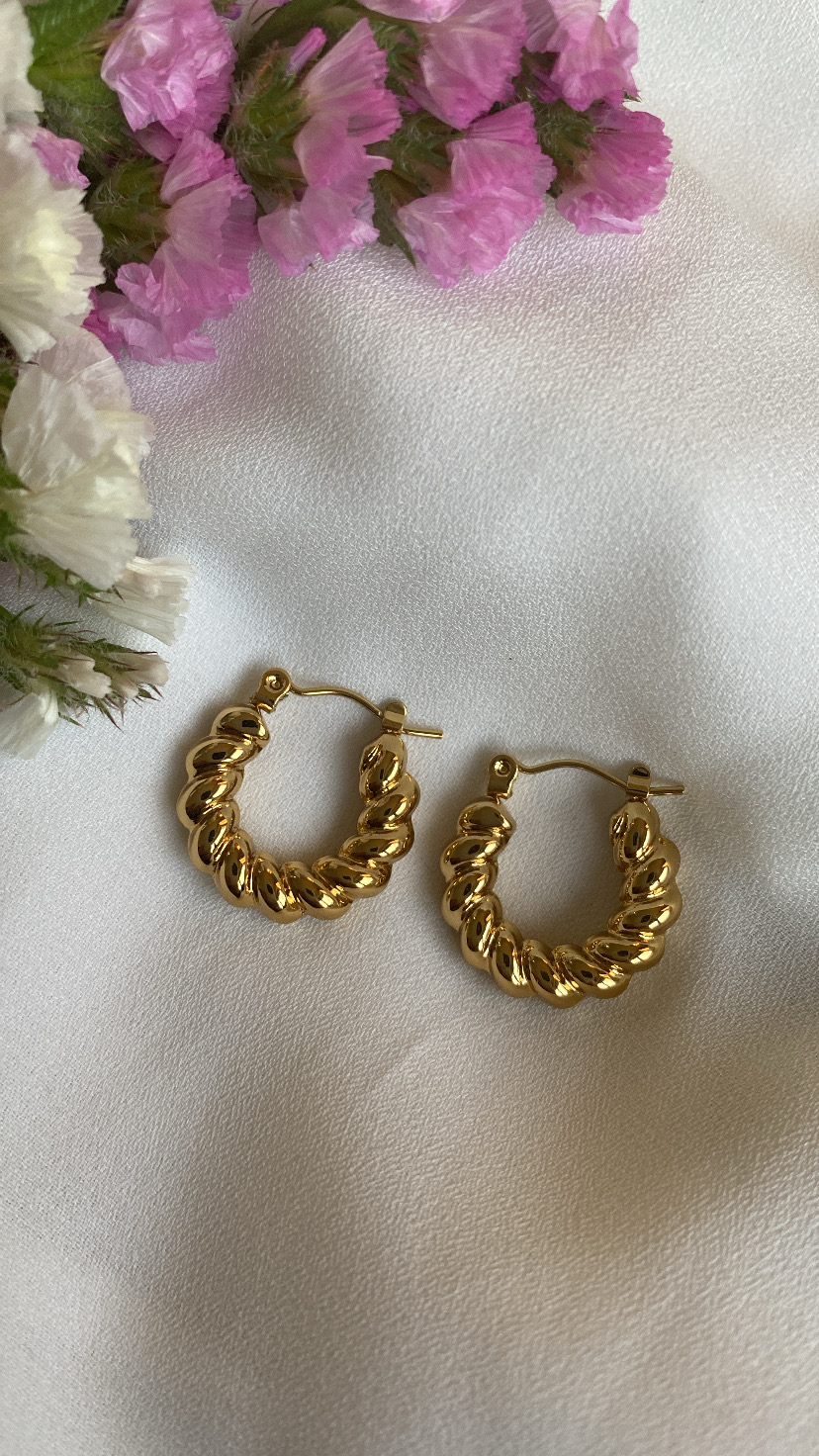 Brincos de argola dourados com textura ondulada sobre tecido branco, com flores rosa e brancas ao lado